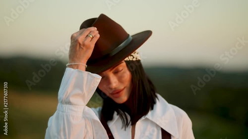 Woman adjusting brown hat in a rural field during the golden sunset hour