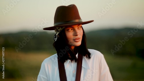 Young woman wearing a stylish brown hat poses in a grassy field during a beautiful golden sunset