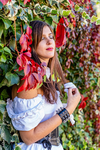 Gothic woman wearing a white dress and gloves, posing among dense ivy and vivid red autumn leaves in a natural outdoor setting, creating a romantic and mysterious fashion scene