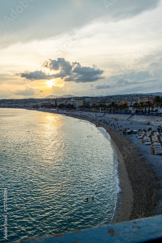 Sunset over Nice, France with calm Mediterranean waters, curved shoreline, beach loungers, palm‑lined promenade, and warm golden light creating a serene coastal atmosphere.
