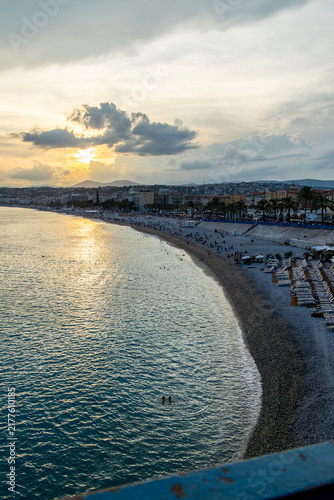 vertical view Sunset over Nice, France with calm Mediterranean waters, curved shoreline, beach loungers, palm‑lined promenade, and warm golden light creating a serene coastal atmosphere.