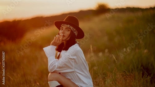 Woman in a hat relaxing in a grassy field at sunset near mountains