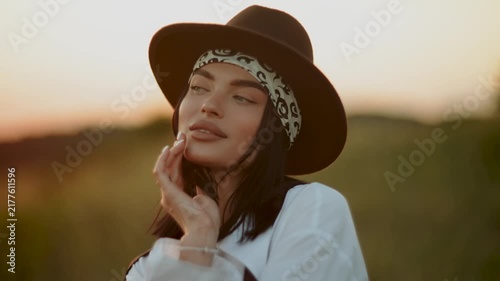 Young woman wearing a brown hat and white shirt standing in a field during a warm golden sunset