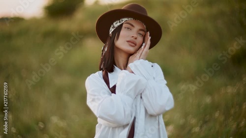 Young woman in a stylish hat posing in a green field during the warm golden sunset hours