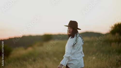 Woman walking through a grassy field at sunset wearing a hat and white shirt