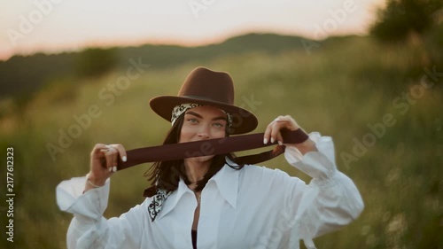 Stylish young woman wearing a brown hat posing in a grassy field during a warm golden sunset