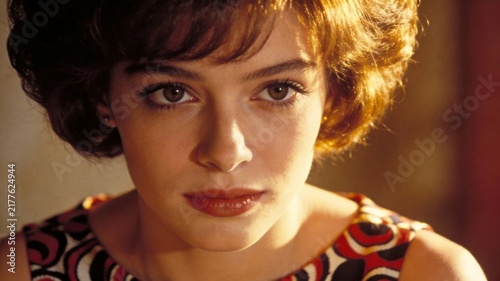 Close-up portrait of a serious young woman with short brunette hair, illuminated by warm cinematic light in a vintage interior.