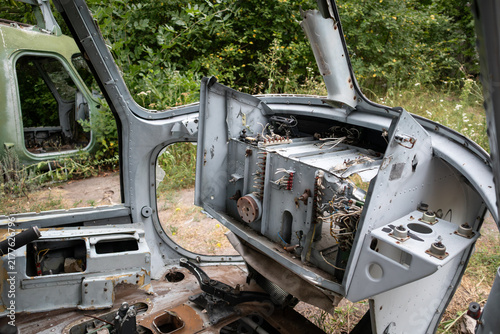 Decayed helicopter cockpit with exposed controls and wiring, surrounded by dense greenery outside the wreck