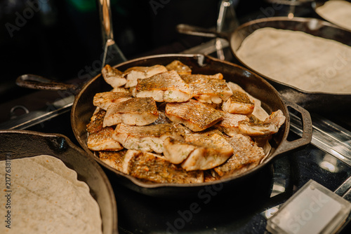 Cooked fish fillets arranged in a cast iron skillet on a buffet table with tortillas and serving utensils visible in the background