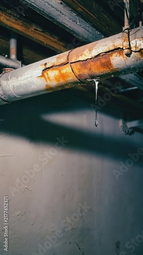 Rusted pipe leaking water, close-up view, showing corrosion, damage, and potential plumbing emergency in a dark, industrial basement setting.