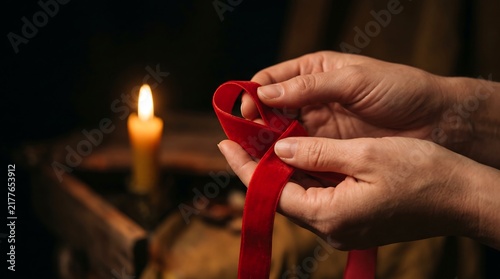 Close-up of hands holding a red ribbon in front of a glowing candle in a dimly lit room, symbolizing awareness and support
