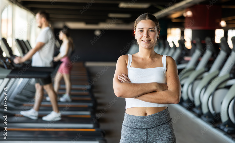Fototapeta premium Young athletic woman in sportswear posing near treadmill in gym