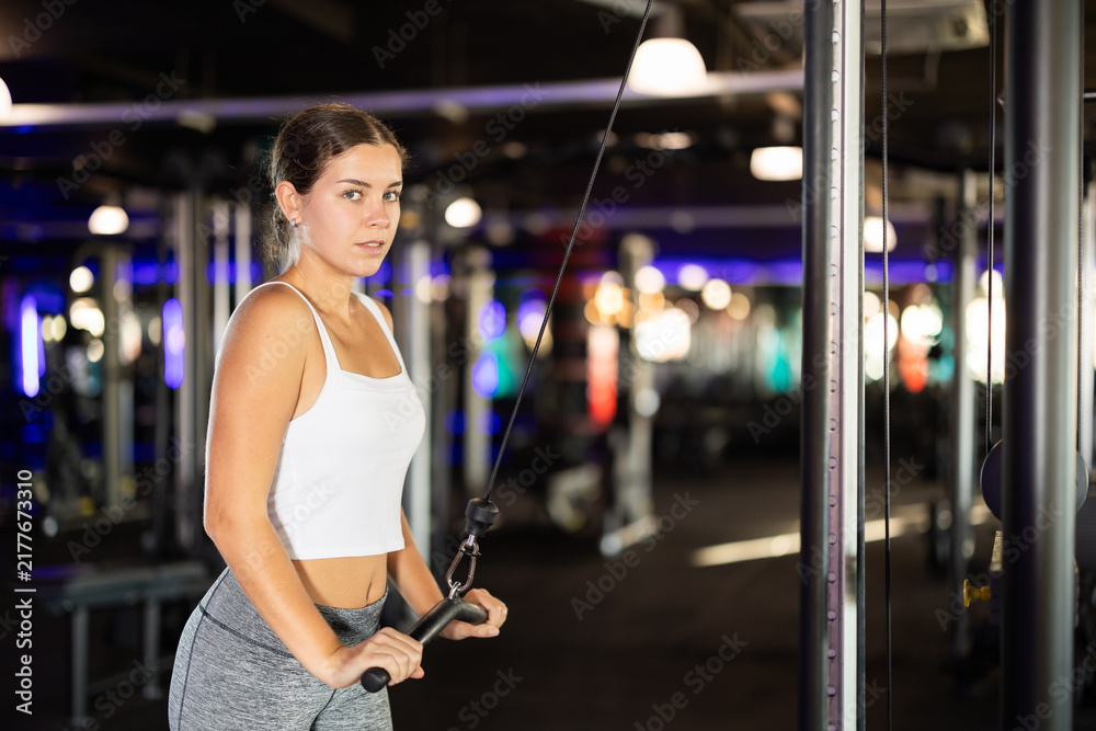 Fototapeta premium Slender athletic girl work out muscles of hands and arms on cable crossover machine in fitness club. Gym visitor trains tris, during classes at multi-station in wellness center.