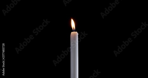Tall taper candle burning steadily in a dark empty room. A bright flame on this isolated white candle cuts through the shadows. The glowing candle symbolizes peace and spiritual warmth.