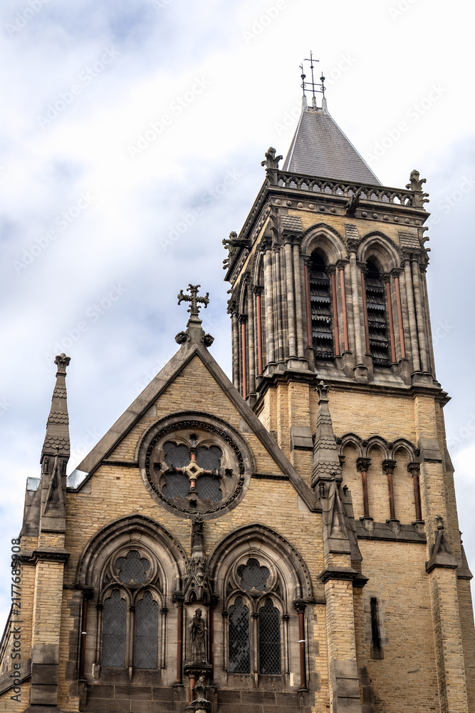 Fototapeta premium Exterior of St Wilfrid’s Church at the York Oratory in York, England, featuring an ornate Gothic Revival facade, bell tower and arched windows beneath a cloudy sky