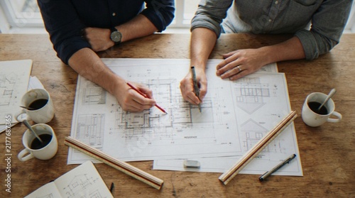 Konstfotografi High angle view of two architects collaborating on house floor plans at a wooden table with coffee cups and drafting tools