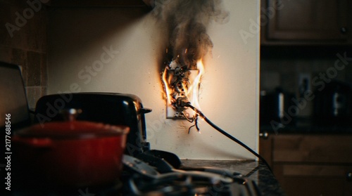 Fotografía An electrical wall outlet catching fire in a kitchen, showing a plug and cord smoking with flames, highlighting a dangerous home hazard