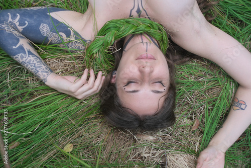 Фотография Tattooed woman resting in green grass with closed eyes and braided plants across