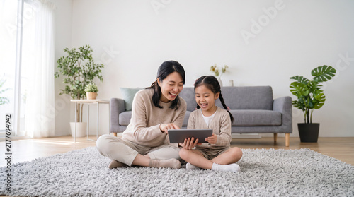 Happy Asian Mother and Little Daughter Laughing While Using Digital Tablet on Living Room Floor