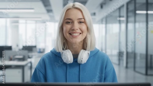 Smiling woman wearing blue hoodie and headphones poses cheerfully in front of office background.