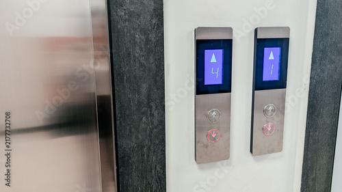Elevator control panel with two buttons. One button is illuminated blue, indicating upward movement. The panel has a sleek, modern design.
