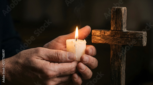 Devout person holding a glowing white candle beside a rustic wooden cross for prayer and spiritual reflection in a dimly lit setting