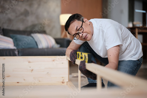 Asian mature man determinedly assembling wooden furniture with DIY tools, checking work precision with spirit level, home improvement, craft installation, hobby carpentry workshop, active lifestyle.