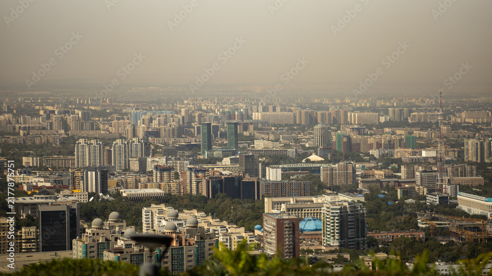 Fototapeta premium Wide panoramic view of Almaty, Kazakhstan, showing a dense urban landscape with modern skyscrapers, residential buildings, business centers, parks and city infrastructure under a hazy sky.