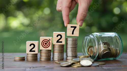 Year 2027 financial goals and savings concept photo. Hand placing wooden block with number seven on a stack of coins next to glass jar on wooden table with a soft green bokeh background