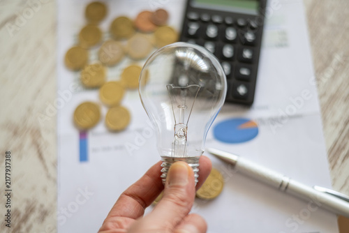 Hand holding an incandescent light bulb over financial documents with coins, a calculator, and a pen, symbolizing rising energy prices and utility expenses