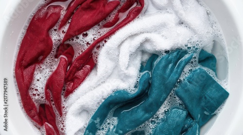 Red, white, and teal fabric clothes soaking in a basin with soapy water and foam bubbles for laundry cleaning