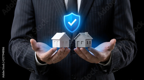 Businessperson in suit holding two 3D rendered house models with a blue shield icon above, professional and secure concept featuring hands, property, real estate, housing, home and checkmark