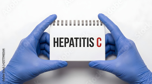 Hands in blue nitrile gloves holding a white spiral-bound notebook with the text HEPATITIS C printed in bold black and red letters against a plain white background featuring medical and healthcare
