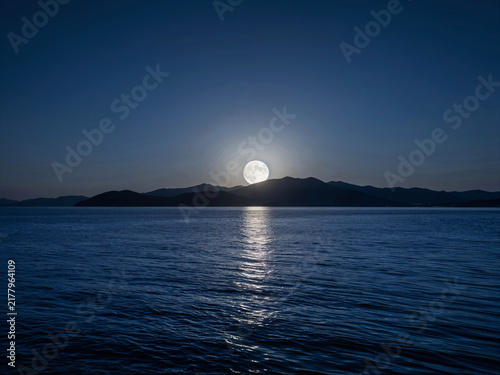 Full moon rising over distant mountains reflecting on a calm lake surf