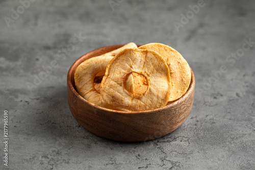 Tasty dehydrated apple chips snack served in small rustic wood bowl on dark concrete background