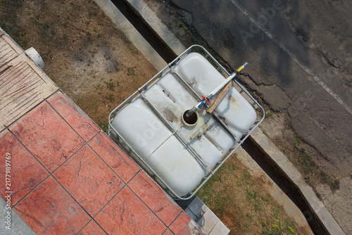 Quadro su tela A high-angle shot shows a large white plastic IBC water tank encased in a metal cage, placed on the ground next to a tiled patio and asphalt driveway