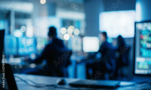 A blurred office scene depicting individuals working at computers in a high-tech environment. Monitors display various data, enhancing the atmosphere of productivity.