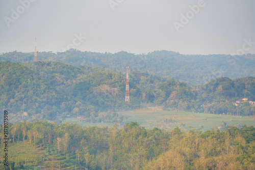 Telecommunication antenna tower standing among lush green hills and rice fields in a hazy countryside landscape under bright sunlight.