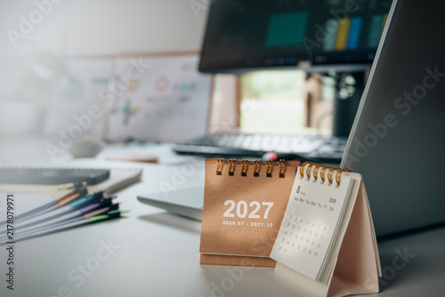 2027 desk calendar on a modern home office workspace with laptop, notebook, stacks of documents and natural window light, representing new year planning, business goals, productivity, future strategy
