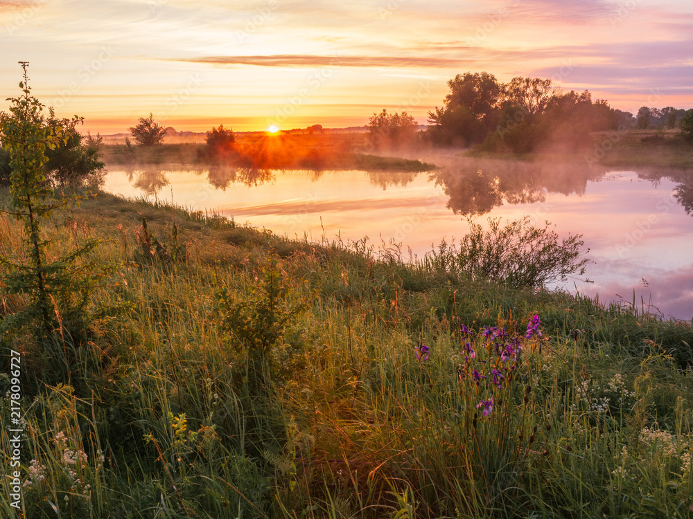 Obraz premium A beautiful misty sunrise illuminates a calm river surrounded by lush green banks and wildflowers