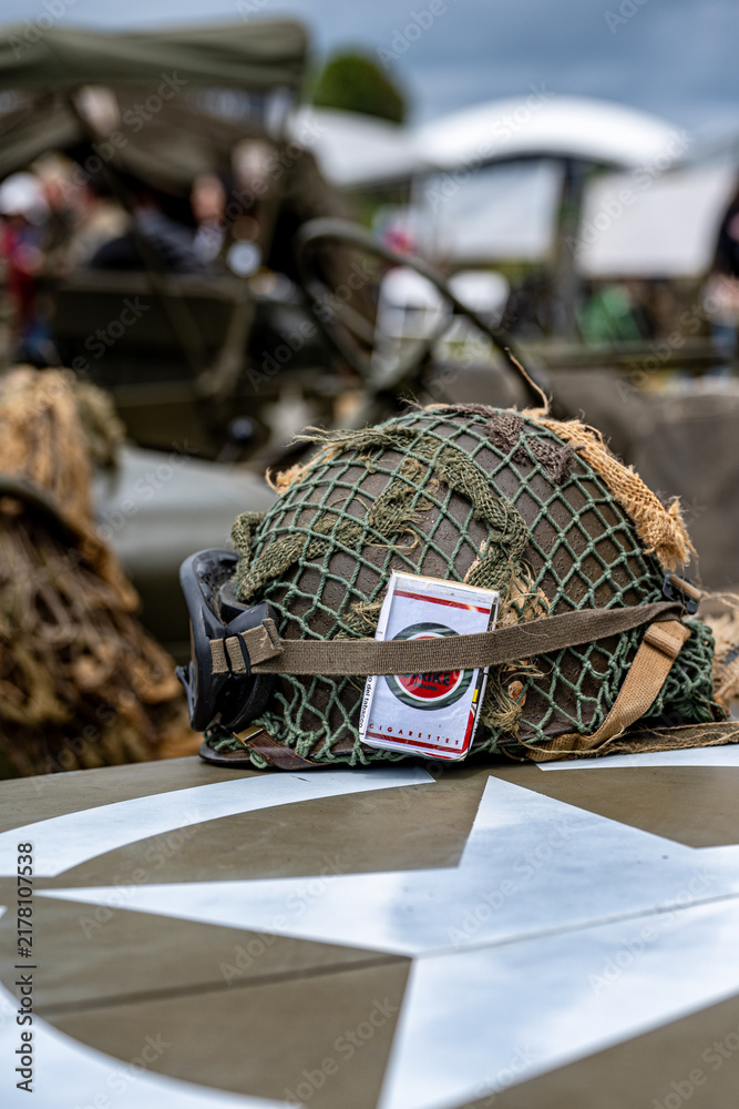 Obraz premium Sainte-Mere-Eglise, France - June 5, 2026: WWII M1 helmet with net and vintage Lucky Strike cigarette pack tucked under the strap on a military vehicle. D-Day 82nd anniversary commemoration at Camp Ge