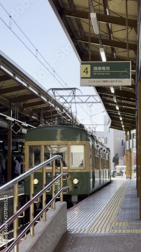 Kamakura, Japan. Green electric train approaches platform at a busy station, with visible signage and passengers waiting along the platform under a clear blue sky
