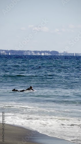 Kamakura, Japan. Surfer paddles through ocean waves on a sunny day, with distant coastline and clear blue sky creating a vibrant beach scene