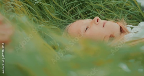 Cinematic side view of a young girl lying in tall grass and looking upward. The shot is filmed through grass in the foreground, creating shallow depth of field and a dreamy mood. High quality 4k