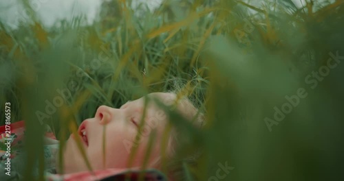 Cinematic view of a young girl lying in green grass and looking upward. Tall grass partially covers the foreground, creating a soft natural frame. Warm natural light and peaceful outdoor atmosphere.