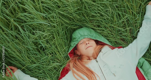 Top-down view of a young girl lying on her back in green grass and looking directly up at the camera. Static overhead shot with natural light and a peaceful outdoor atmosphere. High quality 4k footage