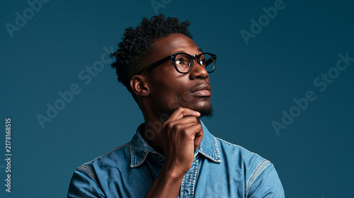 Portrait of pensive african young man wearing denim shirt and eyeglasses, looking up with hand on chin. Studio portrait on blue background