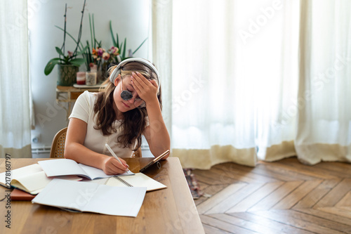 A teenage girl with strabismus wearing corrective eyeglasses with an occluder patch sits at a desk preparing for school. She wears headphones and rests her head while studying.