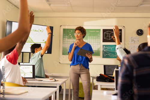 In high school, teacher holding tablet and engaging students raising hands in classroom