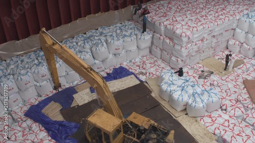 high angle view inside a bulk carrier cargo hold showing a yellow excavator and stevedores neatly stowing tiers of white jumbo cargo bags during loading operations